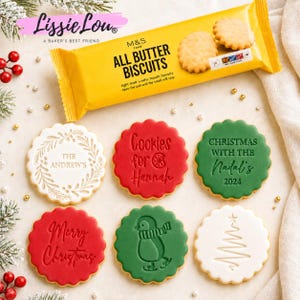 Puede incluir: Un paquete amarillo de galletas M&S Food All Butter Biscuits se sitúa sobre seis galletas redondas de borde festoneado. Las galletas presentan diseños en relieve que incluyen una corona con texto, Cookies for Hannah, Christmas with the Nadal's 2024, Merry Christmas, un pingüino y un árbol.