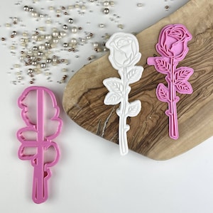 Puede incluir: Tres cortadores de galletas de plástico rosa en forma de rosas. Dos de los cortadores son blancos y uno es rosa.