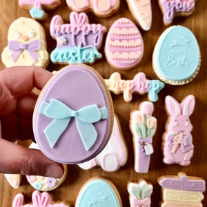 Puede incluir: Una colección de galletas de Pascua decoradas con colores pastel. Las galletas presentan formas como huevos, conejos, pollitos y zanahorias. Algunas galletas tienen el texto "Happy Easter" y "hop hop".