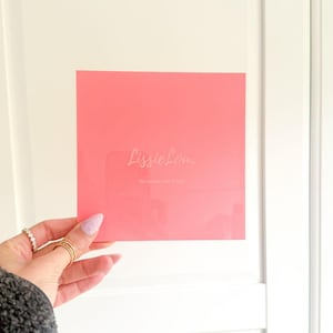 Peut inclure: Une carte carrée rose avec le texte "Lissie Lou" et "6in Square Cake Shops" dans une couleur claire. La carte est tenue par une main avec des bagues et du vernis à ongles rose pailleté.