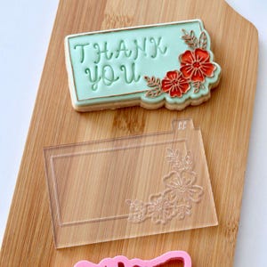 May include: A decorated cookie with the words "THANK YOU" in green script, adorned with red flowers and leaves. A clear plastic cookie stamp with a floral design is also visible, along with a wooden cutting board.