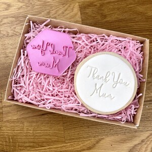 May include: A pink cookie cutter with the text "Thank You Mum" and a white cookie with the same text.