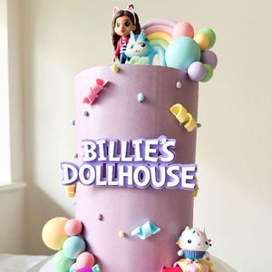 Könnte beinhalten: Ein lila Kuchen, verziert mit Figuren, einem Regenbogen und bunten Süßigkeiten. Der Kuchen trägt den Text "Billie's Dollhouse" in weißen Buchstaben mit lila Umrandung. Der Kuchen ist mit verschiedenen dekorativen Elementen verziert.