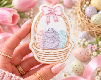 May include: A sugar biscuit shaped like an Easter basket with white icing, featuring a pink piped bow at the top and three decorated eggs in pastel shades of pink, purple, and blue nestled inside the basket outline.