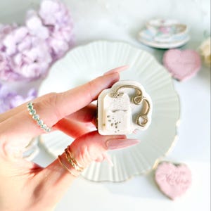 Peut inclure: Une confiserie décorative en forme de sachet de thé blanc avec des détails dorés. La confiserie est tenue dans une main, avec une assiette blanche et une tasse à thé en arrière-plan. Un biscuit en forme de cœur est également visible.