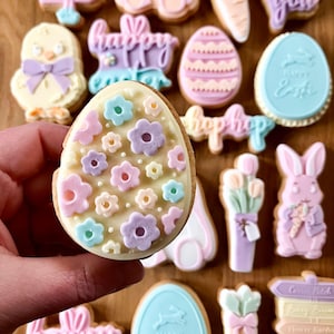 Puede incluir: Una variedad de galletas con temática de Pascua. Las galletas incluyen formas de huevo decoradas con flores, un pollito con un lazo, un conejito y galletas con las palabras "Happy Easter" y "hop hop". Las galletas están decoradas con colores pastel.