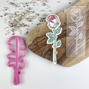 Puede incluir: Un cortador de galletas de plástico rosa con forma de rosa con hojas. Una galleta blanca decorada con glaseado rojo y verde está al lado de una plantilla de plástico transparente de una rosa.