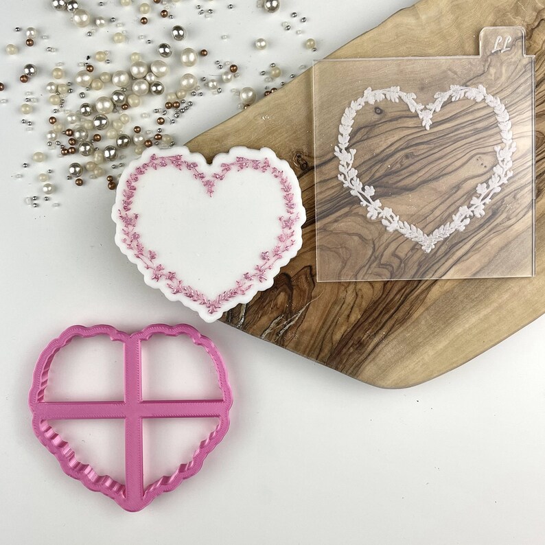 K&ouml;nnte beinhalten: Ein herzf&ouml;rmiger Keks mit rosa floralen Verzierungen, ein rosa herzf&ouml;rmiger Ausstecher und eine transparente Schablone mit einem floralen Herzdesign. Die Artikel befinden sich auf einem Holzbrett mit verstreuten Perlen.