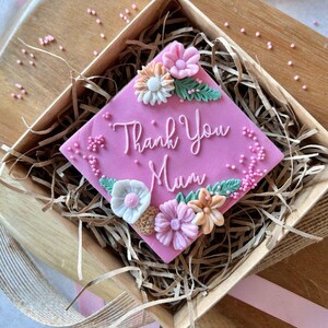 以下が含まれることがあります： ピンクの正方形のケーキに花と「Thank You Mum」の文字が飾られています。