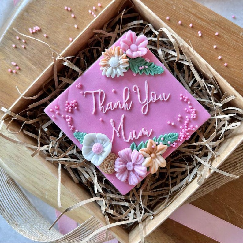 以下が含まれることがあります： ピンクの正方形のケーキに花と「Thank You Mum」の文字が飾られています。