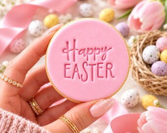 May include: A round sugar cookie with smooth pink icing featuring the embossed text Happy EASTER. The cookie is held against a background of pink tulips, a small woven nest filled with speckled pastel eggs, and pink ribbon.