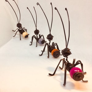 Puede incluir: Una fila de hormigas de arte de alambre hechas a mano. Cada hormiga tiene un cuerpo, patas y antenas de alambre marr&oacute;n oscuro. Las hormigas tienen coloridos abd&oacute;menes de cuentas en tonos de rosa, naranja, lavanda y crema. Las hormigas est&aacute;n dispuestas en diagonal.
