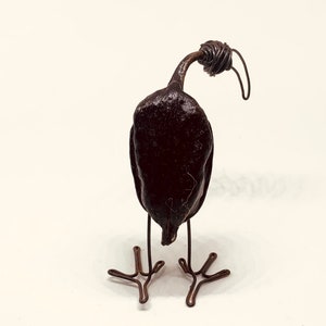 Puede incluir: Una pequeña escultura de pájaro negra en forma de pájaro, hecha de una calabaza seca y alambre. El pájaro tiene un cuello largo y una cabeza de alambre con un detalle de alambre rizado. El pájaro está de pie sobre dos patas de alambre con tres dedos de alambre en cada pie.