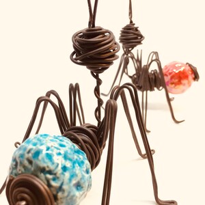 May include: Two wire sculptures of ants with ceramic heads. The ant on the left has a blue and white head, while the ant on the right has a pink and white head.