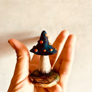 Puede incluir: Una peque&ntilde;a figurita de hongo de cer&aacute;mica pintada a mano con un sombrero azul y puntos naranjas. El hongo est&aacute; sentado sobre una base de madera con detalles verdes.