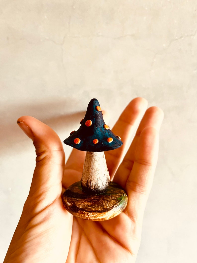 Puede incluir: Una peque&ntilde;a figurita de hongo de cer&aacute;mica pintada a mano con un sombrero azul y puntos naranjas. El hongo est&aacute; sentado sobre una base de madera con detalles verdes.
