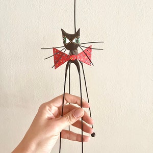 Puede incluir: Una figurita de gato negro con una pajarita roja y ojos verdes. El gato tiene patas largas y delgadas y una cola larga. La figurita est&aacute; hecha de metal y tiene un estilo caprichoso y de dibujos animados.