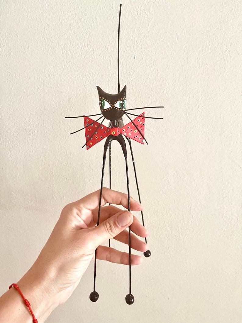 Puede incluir: Una figurita de gato negro con una pajarita roja y ojos verdes. El gato tiene patas largas y delgadas y una cola larga. La figurita est&aacute; hecha de metal y tiene un estilo caprichoso y de dibujos animados.