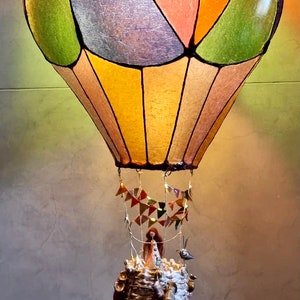 Może przedstawiać: Dekoracyjna lampa w kształcie balonu na ogrzane powietrze z wielokolorowym kloszem. Balon ma zielone, pomarańczowe i fioletowe panele. Poniżej wisi kosz z drabiną i małymi figurkami. Mały ptaszek siedzi na g&oacute;rze.