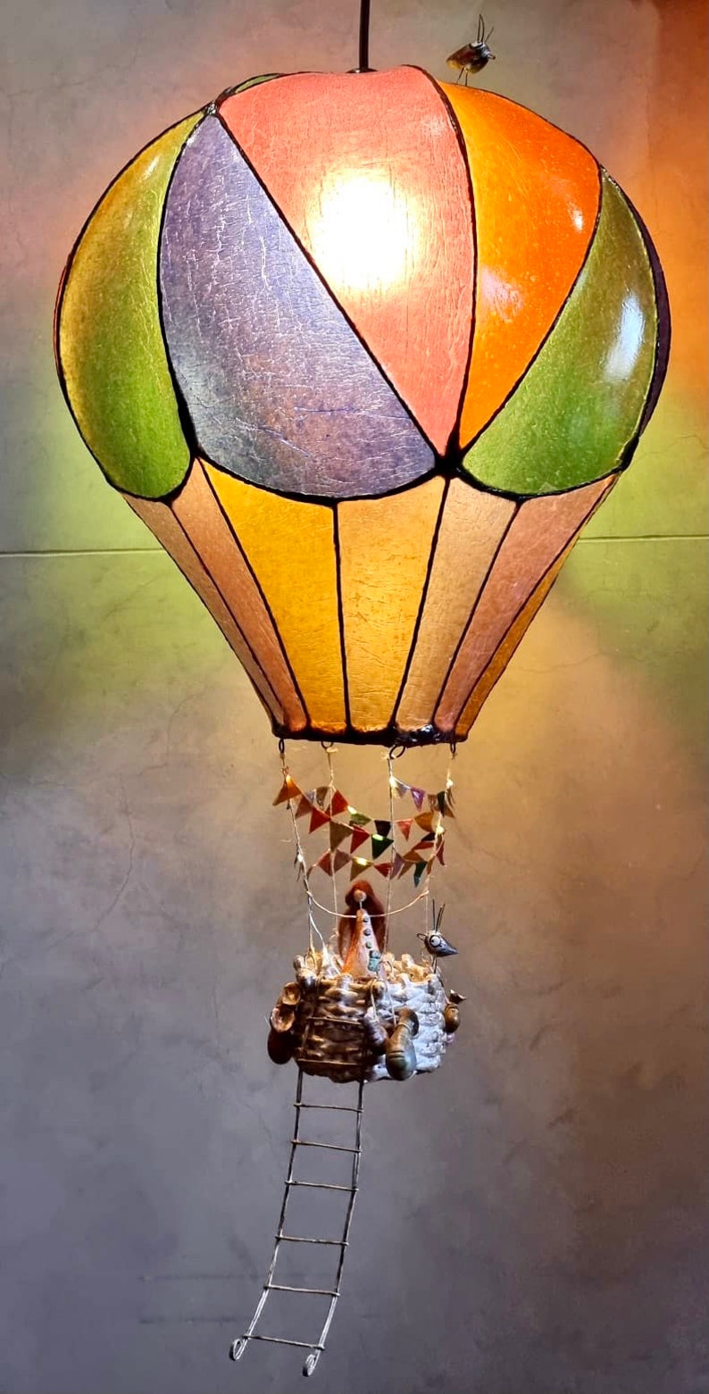 Może przedstawiać: Dekoracyjna lampa w kształcie balonu na ogrzane powietrze z wielokolorowym kloszem. Balon ma zielone, pomarańczowe i fioletowe panele. Poniżej wisi kosz z drabiną i małymi figurkami. Mały ptaszek siedzi na g&oacute;rze.