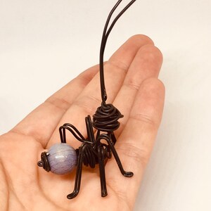 Puede incluir: Una escultura de alambre artesanal de una hormiga. La hormiga est&aacute; hecha de alambre negro con una cuenta esf&eacute;rica de color lavanda para la cabeza. Las antenas tienen extremos en bucle. La escultura se sostiene en una mano.