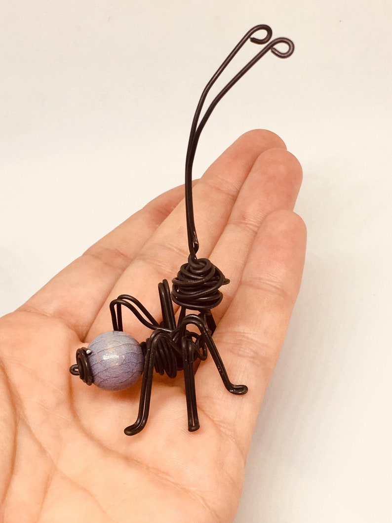 Puede incluir: Una escultura de alambre artesanal de una hormiga. La hormiga est&aacute; hecha de alambre negro con una cuenta esf&eacute;rica de color lavanda para la cabeza. Las antenas tienen extremos en bucle. La escultura se sostiene en una mano.