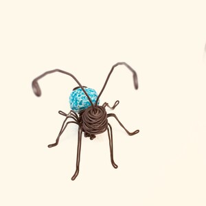 May include: A wire sculpture of an ant with a blue and white bead for a head. The ant is made of brown wire and has six legs.