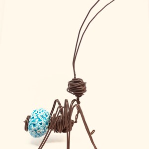 May include: A wire sculpture of an ant with a blue and white ceramic head. The ant is made of brown wire and has long, thin legs. The ant is standing on a white surface.