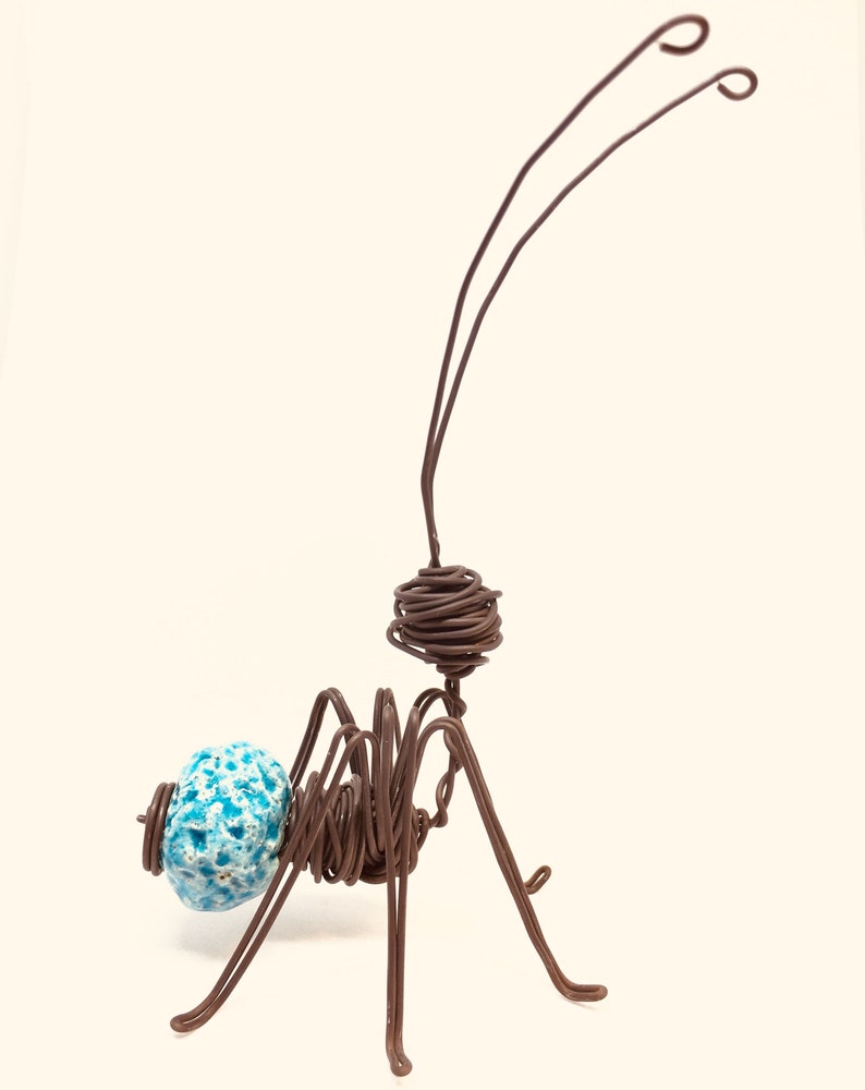 May include: A wire sculpture of an ant with a blue and white ceramic head. The ant is made of brown wire and has long, thin legs. The ant is standing on a white surface.