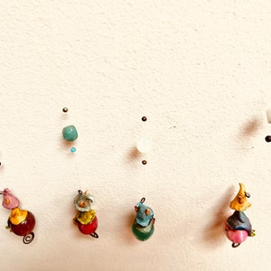 May include: Five colorful ceramic figurines hanging from the ceiling. Each figurine is wearing a hat and has a round body. The figurines are suspended from a wire with beads and other decorative elements.