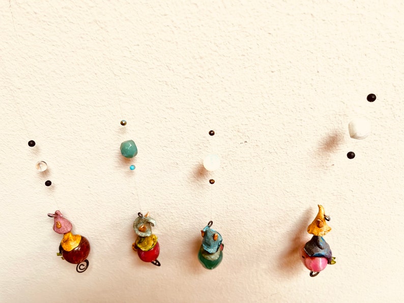 May include: Five colorful ceramic figurines hanging from the ceiling. Each figurine is wearing a hat and has a round body. The figurines are suspended from a wire with beads and other decorative elements.