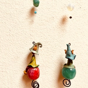 May include: Two handmade ornaments with a whimsical design. Each ornament features a small gnome-like figure with a hat and a round, colorful stone. The ornament on the left has a red stone, while the ornament on the right has a green stone.