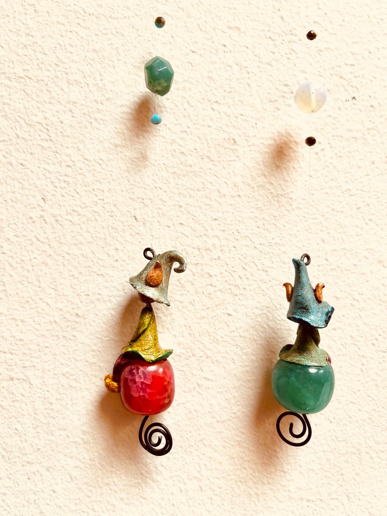 May include: Two handmade ornaments with a whimsical design. Each ornament features a small gnome-like figure with a hat and a round, colorful stone. The ornament on the left has a red stone, while the ornament on the right has a green stone.