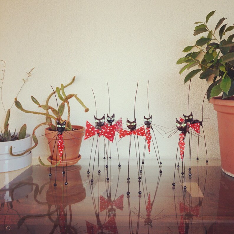 Puede incluir: Un grupo de siete figuras de gato de metal negro con pajaritas de lunares rojos y blancos est&aacute;n de pie sobre una mesa. Los gatos est&aacute;n dispuestos en una l&iacute;nea, con sus colas apuntando hacia arriba. Los gatos est&aacute;n todos mirando hacia adelante, y sus ojos est&aacute;n cerrados. Los gatos llevan todos pajaritas de lunares rojos y blancos. Los gatos est&aacute;n todos de pie sobre una mesa, y hay plantas en el fondo.