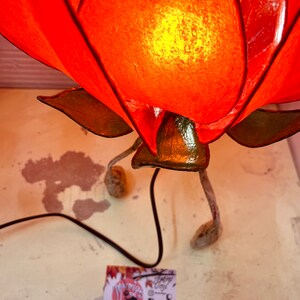 Puede incluir: Una l&aacute;mpara de papel con forma de flor roja con un tallo verde y tres patas. La l&aacute;mpara est&aacute; encendida y la luz brilla a trav&eacute;s del papel.