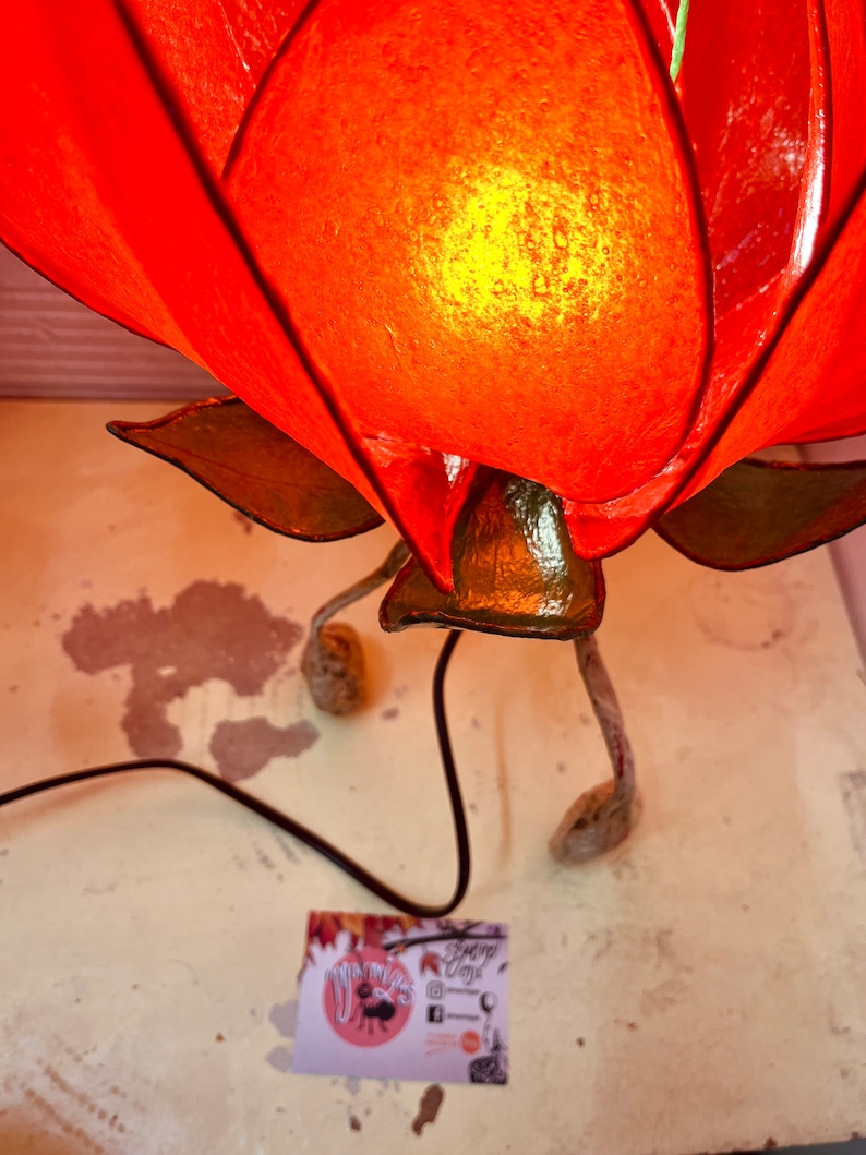 Puede incluir: Una l&aacute;mpara de papel con forma de flor roja con un tallo verde y tres patas. La l&aacute;mpara est&aacute; encendida y la luz brilla a trav&eacute;s del papel.