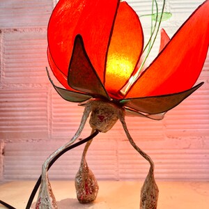 Puede incluir: Una l&aacute;mpara de flores de papel roja con tallo y hojas verdes. La l&aacute;mpara tiene una base blanca con tres patas que se asemejan a capullos de flores.