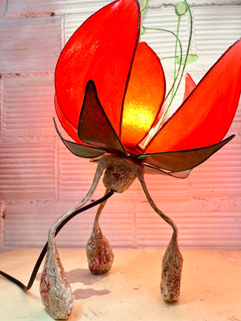 Puede incluir: Una l&aacute;mpara de flores de papel roja con tallo y hojas verdes. La l&aacute;mpara tiene una base blanca con tres patas que se asemejan a capullos de flores.