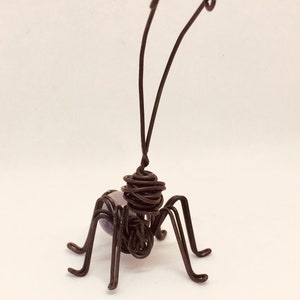 Puede incluir: Una escultura de alambre hecha a mano de un insecto con un acabado marr&oacute;n oscuro. El insecto tiene seis patas, dos antenas y un cuerpo esf&eacute;rico con una cuenta p&uacute;rpura. La escultura est&aacute; sobre un fondo blanco.