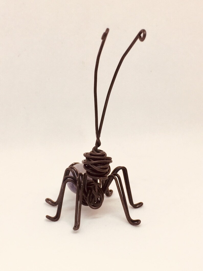 Puede incluir: Una escultura de alambre hecha a mano de un insecto con un acabado marr&oacute;n oscuro. El insecto tiene seis patas, dos antenas y un cuerpo esf&eacute;rico con una cuenta p&uacute;rpura. La escultura est&aacute; sobre un fondo blanco.