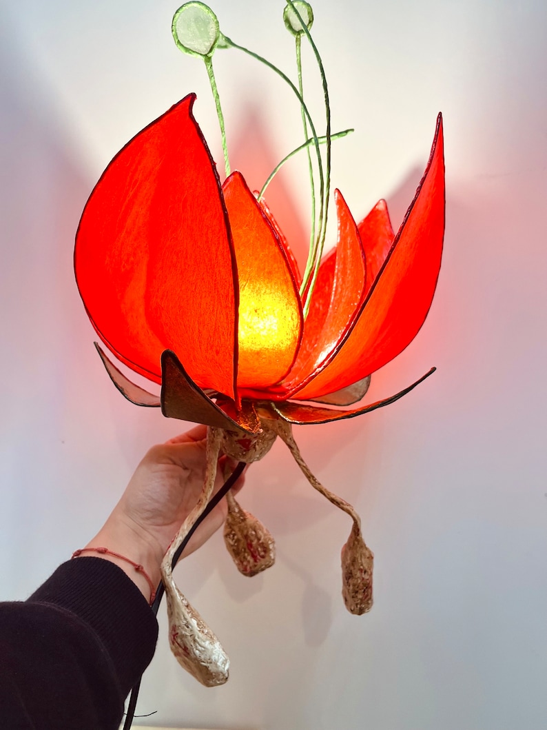 Puede incluir: Una l&aacute;mpara de papel con forma de flor roja con una luz en su interior. La l&aacute;mpara tiene un cable largo y tres tallos hechos de papel con un acabado dorado.