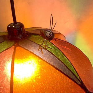 Może przedstawiać: Kolorowa lampa wisząca ze witrażu z dekoracyjną figurką owada. Lampa posiada okrągły, segmentowy klosz w odcieniach pomarańczu, zieleni i brązu. Owad siedzi na kloszu, dodając fantazyjnego akcentu do oprawy oświetleniowej.