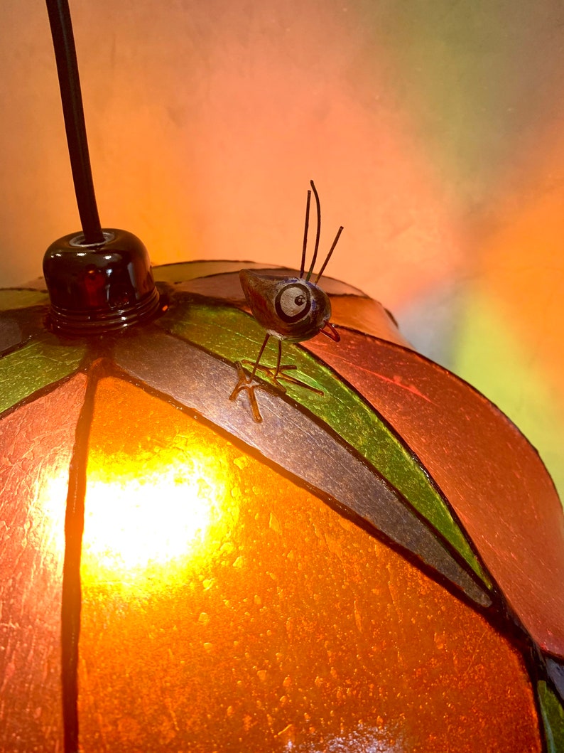 Może przedstawiać: Kolorowa lampa wisząca ze witrażu z dekoracyjną figurką owada. Lampa posiada okrągły, segmentowy klosz w odcieniach pomarańczu, zieleni i brązu. Owad siedzi na kloszu, dodając fantazyjnego akcentu do oprawy oświetleniowej.
