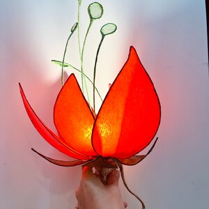 Puede incluir: Una l&aacute;mpara de papel con forma de flor roja con tallos y hojas verdes. La l&aacute;mpara est&aacute; encendida y emite un brillo c&aacute;lido.