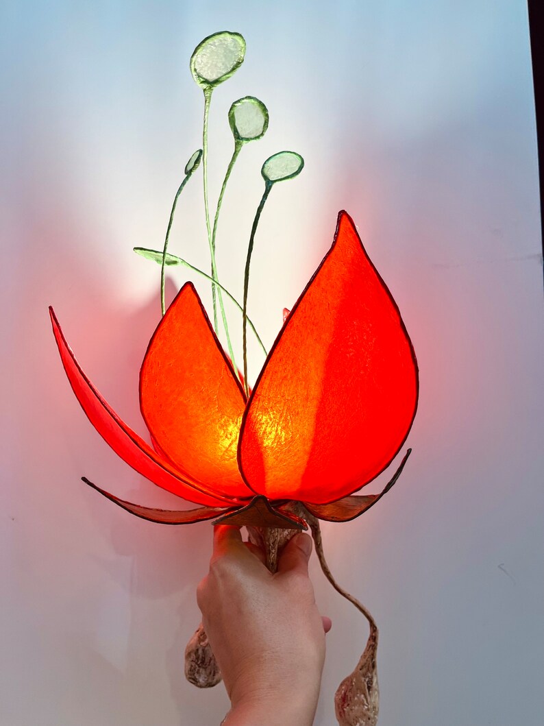 Puede incluir: Una l&aacute;mpara de papel con forma de flor roja con tallos y hojas verdes. La l&aacute;mpara est&aacute; encendida y emite un brillo c&aacute;lido.