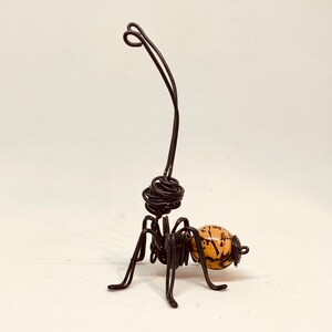 Puede incluir: Una escultura de alambre hecha a mano de un insecto con una antena larga y curvada. El cuerpo del insecto est&aacute; hecho de alambre negro, con una cuenta esf&eacute;rica de color beige con marcas marr&oacute;n oscuro. La escultura tiene seis patas y una cabeza enrollada.
