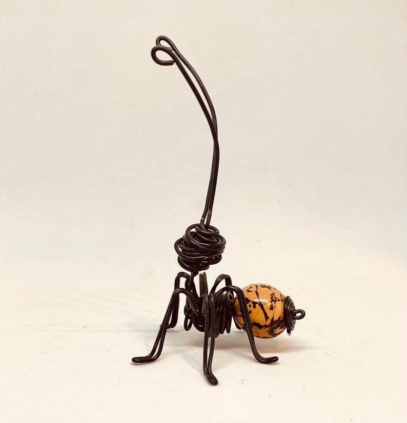 Puede incluir: Una escultura de alambre hecha a mano de un insecto con una antena larga y curvada. El cuerpo del insecto est&aacute; hecho de alambre negro, con una cuenta esf&eacute;rica de color beige con marcas marr&oacute;n oscuro. La escultura tiene seis patas y una cabeza enrollada.