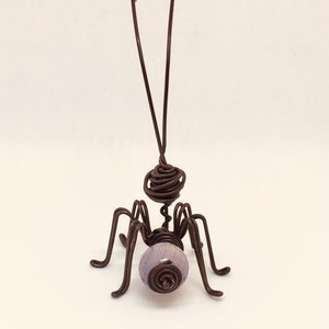 Puede incluir: Una escultura de insecto hecha a mano con alambre. El insecto tiene una cabeza y un cuerpo enrollados, seis patas y dos antenas. Una cuenta de color lavanda forma el abdomen. La escultura est&aacute; hecha de alambre marr&oacute;n oscuro.