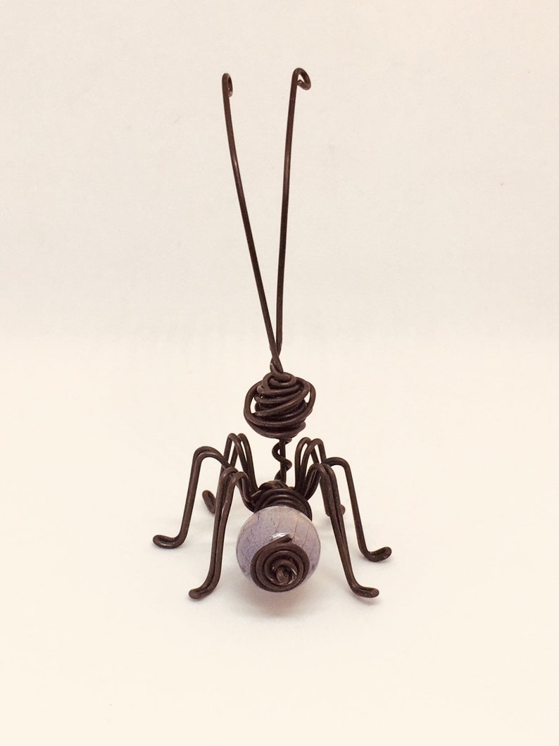 Puede incluir: Una escultura de insecto hecha a mano con alambre. El insecto tiene una cabeza y un cuerpo enrollados, seis patas y dos antenas. Una cuenta de color lavanda forma el abdomen. La escultura est&aacute; hecha de alambre marr&oacute;n oscuro.