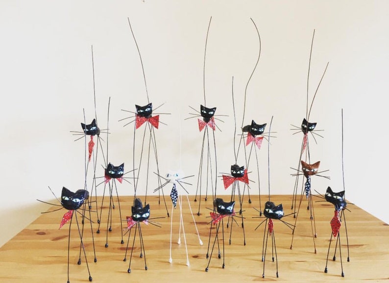 Puede incluir: Una colecci&oacute;n de figuritas de gatos negros con pajaritas rojas y patas largas y delgadas de alambre. Los gatos est&aacute;n dispuestos sobre una superficie de madera.
